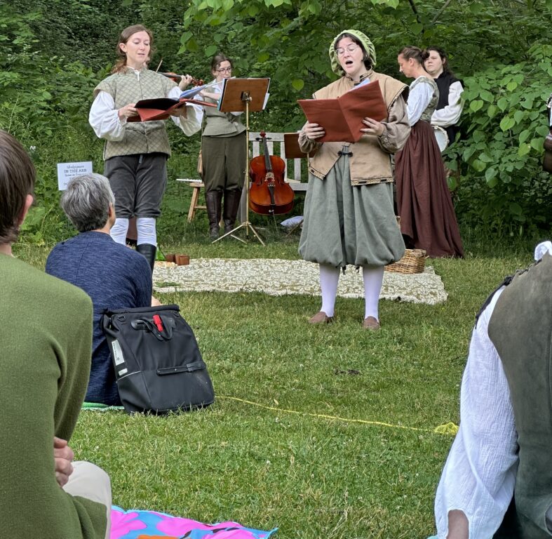 Several musicians and an actor stand outside performing in Shakespearian attire while people sit on the grass in front and watch.