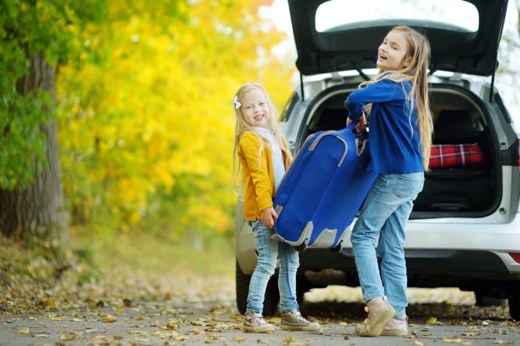 Two girls work together to load a heavy suitcase into the trunk of a car. It's a sunny autumn day.