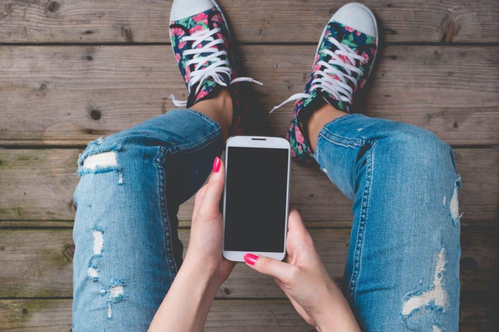 A sitting woman wearing stylishly ripped jeans holds a smartphone in both hands. She's wearing floral-print sneakers. You can only see her hands and legs in the picture.