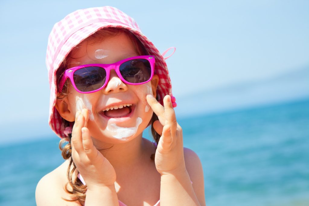 A smiling young girl with a pink hat and pink-rimmed sunglasses applies sunscreen to her face. The ocean is blurred in the background.