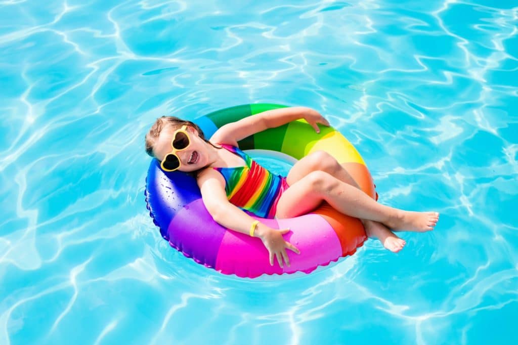 A young girl with heart-shaped sunglasses laughs and smiles on a multi-colored innertube pool toy, floating on clear blue water.