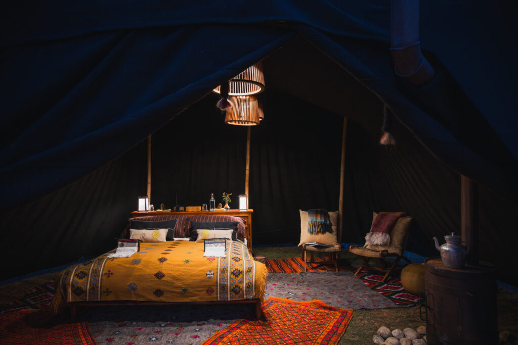 Inside a large tent at night, lit by overhead lamp, are a double bed, two chairs with ottomans and a wood stove, with colorful linens and rugs.