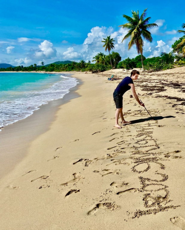 Young teenager uses long stick to write a message in the sand next to the ocean.
