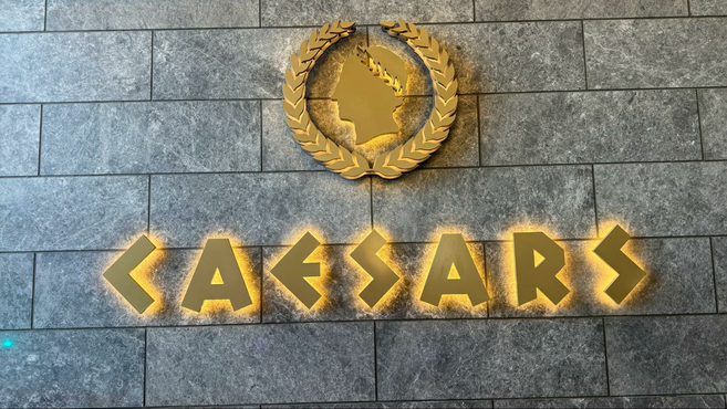 Yellow backlit Caesar's logo against a gray brick wall.