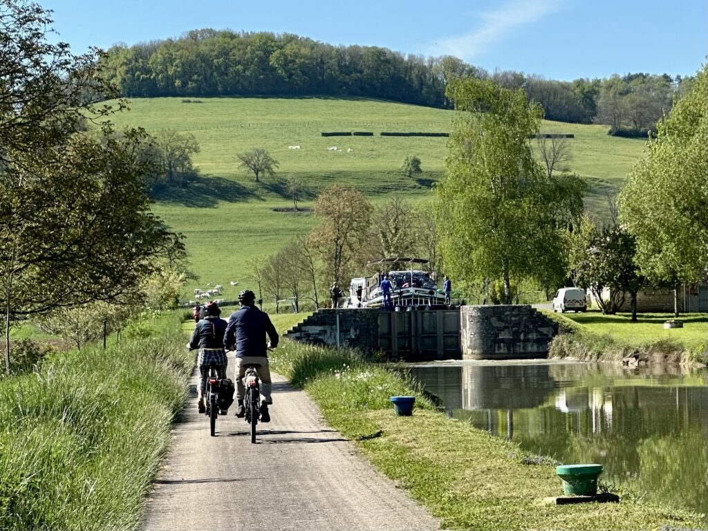 Two cyclists on towpath with a European Waterways hotel barge passing beneath a bridge in Burgundy.