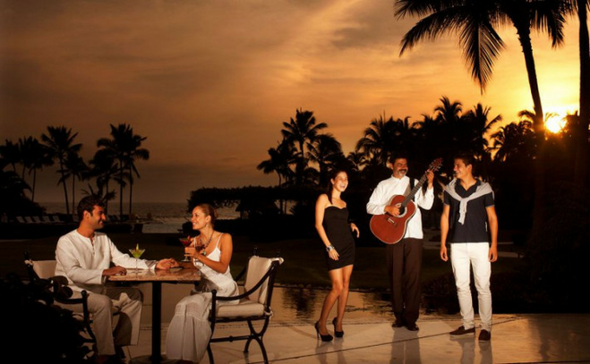Guitarist posing with guests on the main terrace, with a sunset overhead. Nearby, a couple enjoys cocktails at a table