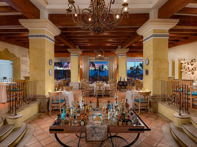 Frida dining room. Just ahead is a cart filled with alcohol spirits. In the background are white-clothed tables ready for dinner service, and elongated columns supporting the ceiling