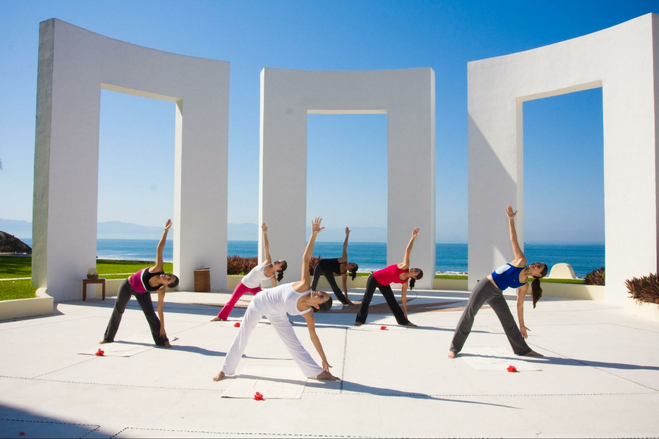 Yoga students holding a pose outside the courtyard. Behind them are three white, towering arches, and a view of the ocean, with cloudless skies above.