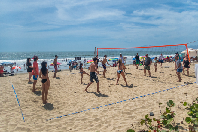 Beach volleyball players enjoying a game, with a volleyball net separating teams. The ocean is in the background