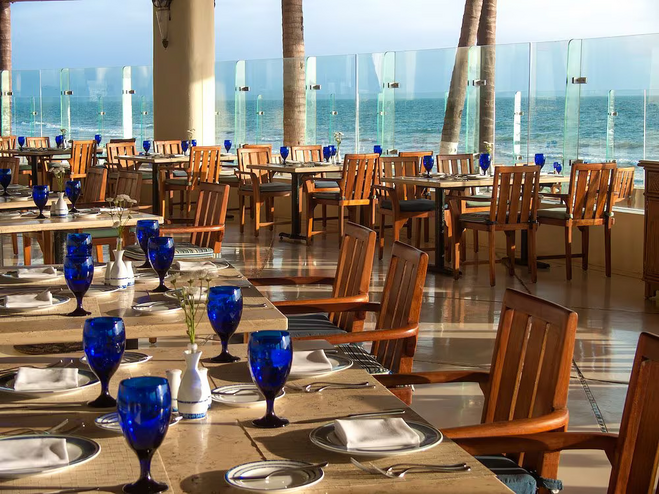 Rows of tables set up for breakfast service, and clear glass windows align the restaurant, with the ocean in the background