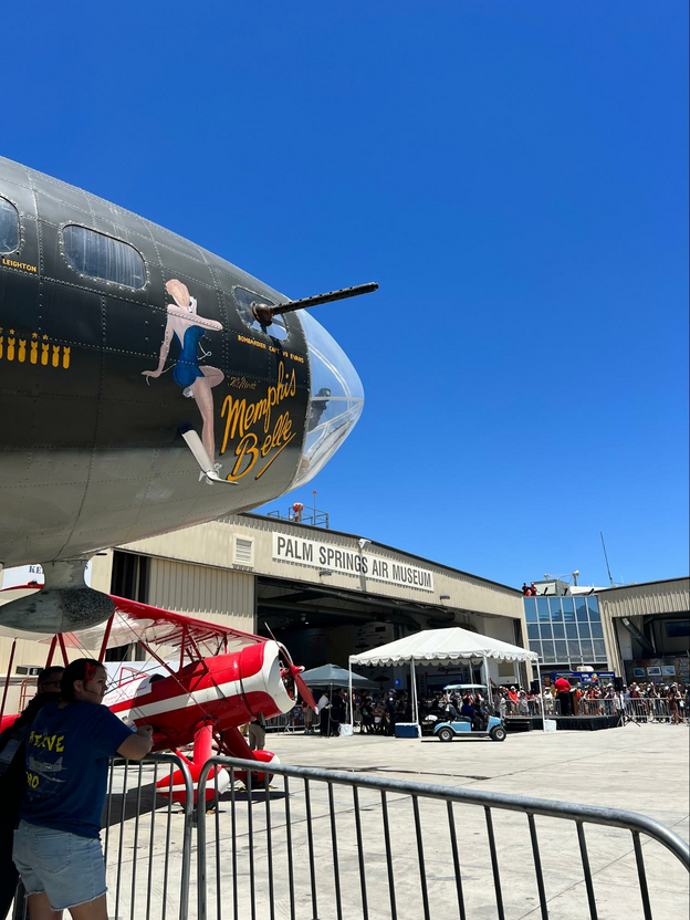 The nose of the Memphis Belle aircraft with the Palm Springs Air Museum behind it and two people standing below the aircraft. A red and white  biplane is between the airplane and the building.