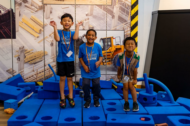 3 children standing in front of picture with construction and a piece of heavy construction equipment. The children are standing on firm blue blocks with holes in them