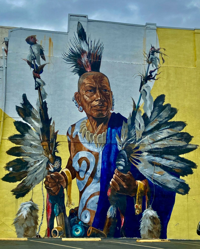 Outdoor building mural of an early native American wearing brightly colored tunic and carrying feathered hunting gear.