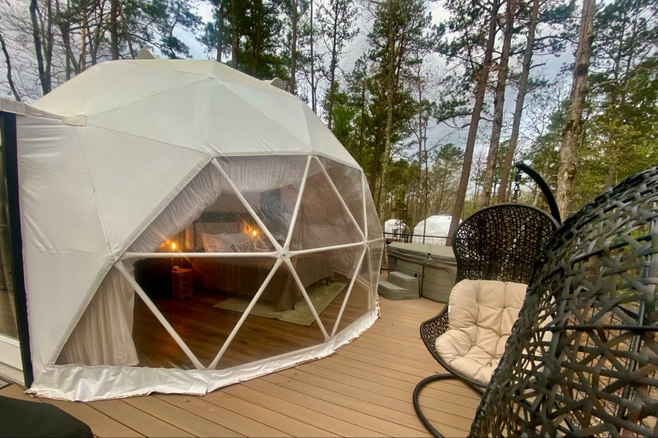 A round tent with a see-through plastic front supported by narrow white rods sitting on a deck with a hot tub and swinging chairs.