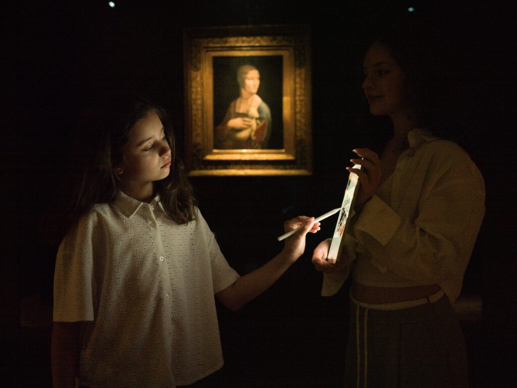 Two young girls painting a digital self-portrait in front of a painting on the wall.