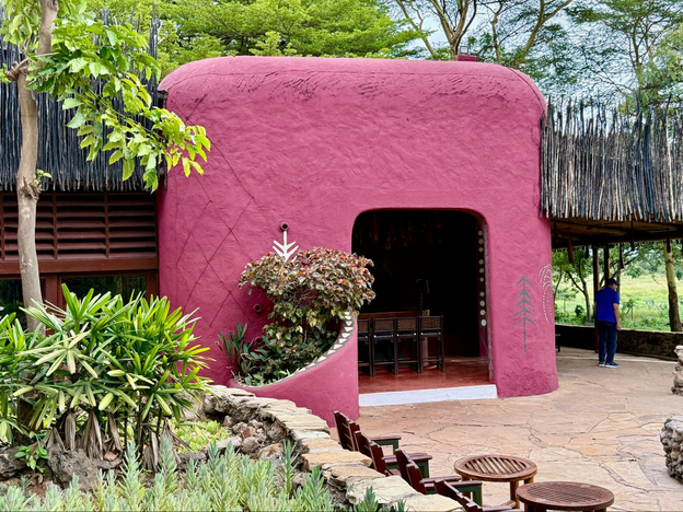 Reddish-pink building with rounded top and smooth walls is flanked with sharp pointed long sticks above head-level. The lodging huts are surrounded with lush trees and gardens/