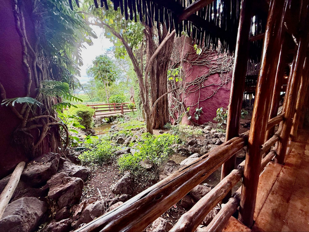 Dark wood walkway with split-rail support beams opens to a glimpse of the reddish-pink smooth-walled lodging buildingsand to dense forest and garden with green bushes amidst big boulders.