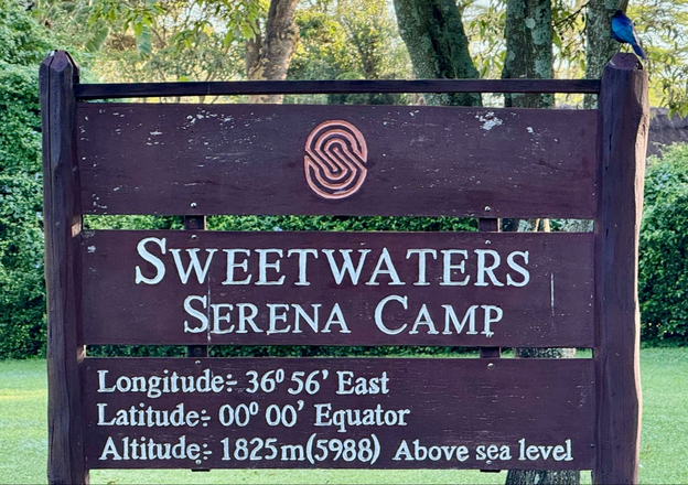 Simple wooden sign with three horizontal boards declaring Sweetwaters Serena Camp at Latitude 00 degrees 00 feet at the Equator. 