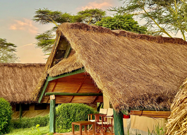 Clouds dot the blue-gray sky and slender green trees stretch taller than the thatch roofs covering spacious lodging tents. Two canvas folding chairs and a small table fill the back porch.