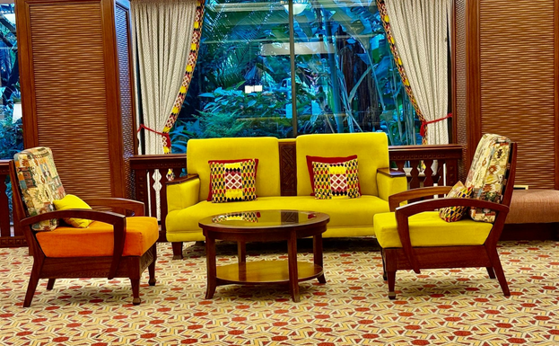 Bold yellow sofa cushions are framed by a large window overlooking dense greenery in a hotel courtyard. Side chairs of the same yellow and also deep orange sit on either side of a round wooden table.