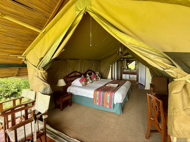 Khaki-yellow tent roof and flaps open to double bed with crisp white sheet. Pillows and coverlet are stripes and diamond shapes in turquoise, red and purple. The sink and mirror are visible with the toilet and shower off to each side. A roof of thatch and sticks covers the tent canvas and extends to the sides.