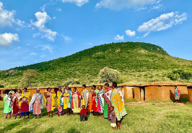 Kenya blue skies with layers of clouds in narrow white wisps reach behind a lush green mountain in a Maasai village. Women in colorful shukas--soft cloths tied around their shoulders-sing in a line in front of their modest square homes.