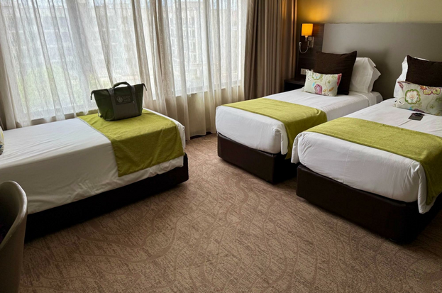 Three twin-size beds in a single room, with a wall of curtained windows and three pillows on each bed.