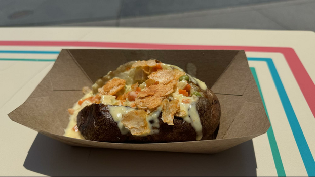 Baked potato stuffed with vegetables, creamy sauce and pie crumbles on top in a cardboard container.