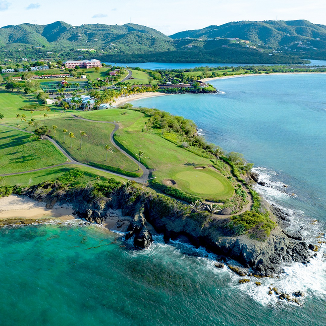 aerial view of golf course showing blue water, a green golf course with resort in the background