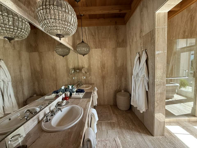 Luxurious tiled Bathroom with ornate crystal light fixtures and two bathrobes hanging behind the vanity area with a view of the tub in the next room