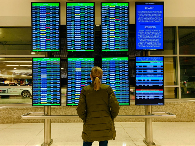 Back of woman in a puffy coat and ponytail and she stands in front of airline monitors showing departures and arrivals.