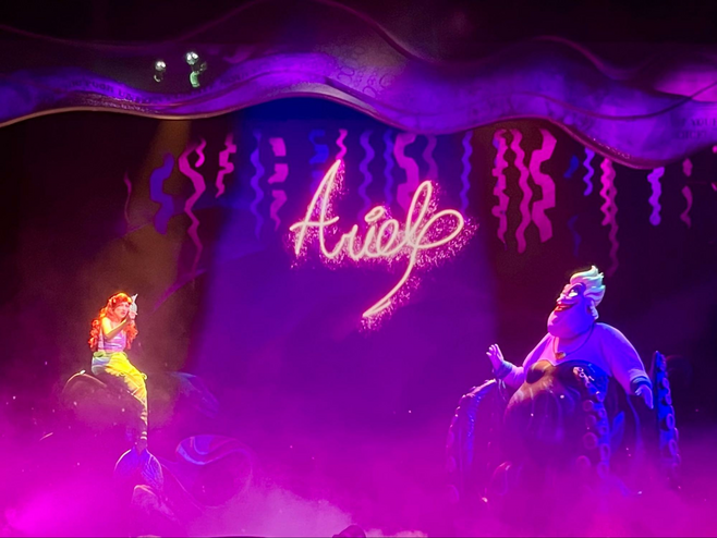 The mermaid Ariel from Disney's The Little Mermaid perches on a rock underwater while signing a contract created by the sea witch, Ursula, as Ursula looks on. Ariel's signature is projected on a screen above them.