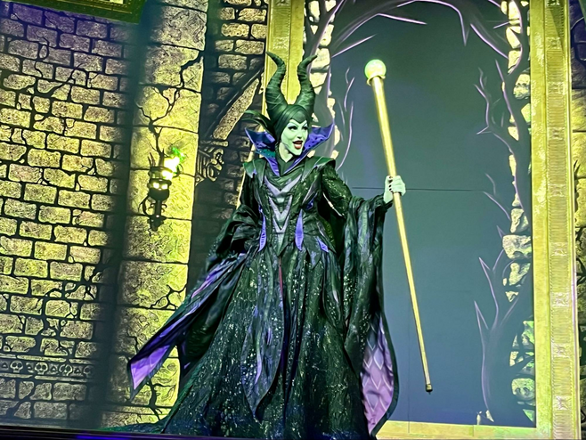 A costumed Maleficent from Disney's Sleeping Beauty is dressed in a black cloaked dress and holds a large golden scepter. Behind her, projections create the image of her castle.