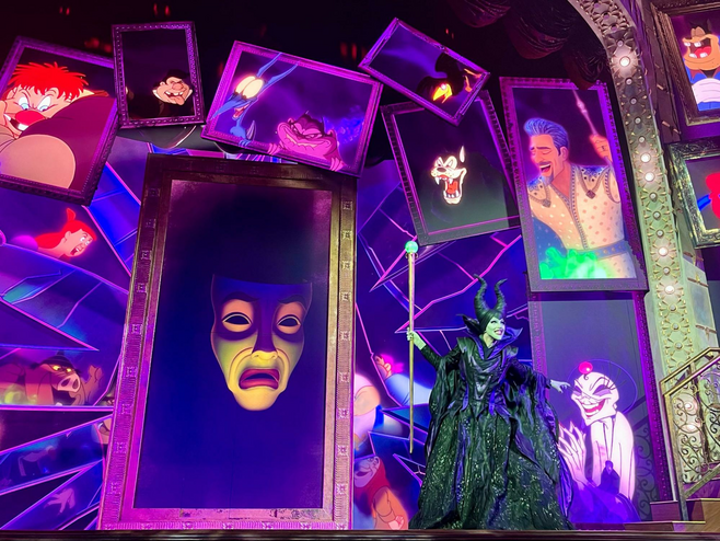 A costumed Maleficent from Disney's Sleeping Beauty is dressed in a black cloaked dress and holds a large golden scepter, smiling at the audience. Behind and above her, images of other Disney villains are projected onto screens that appear as mirrors.