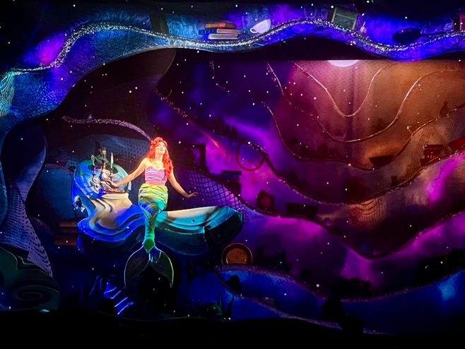 The mermaid Ariel from Disney's The Little Mermaid perches on a rock in her underwater cavern, singing, with rainbow colors of light projected around her.