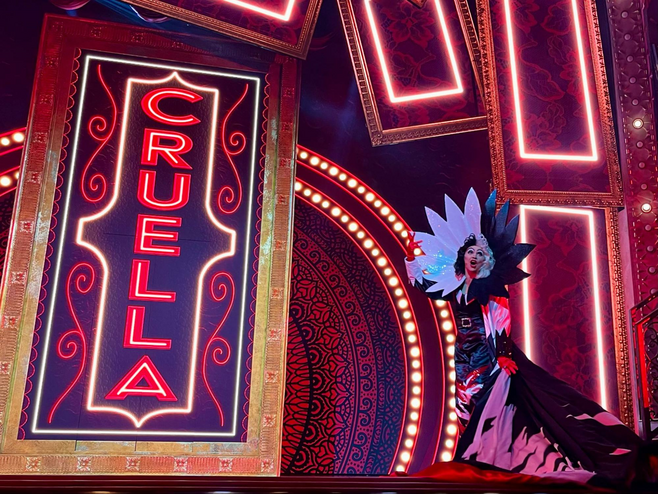 A black-and-white costumed Cruella de Vil sings to audiences next to a huge sign that reads "Cruella."