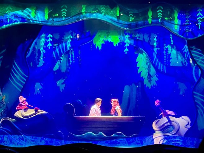 Prince Eric and Ariel from Disney's The Little Mermaid sit in a rowboat onstage with beautiful blue lighting around them during a performance of the song "Kiss the Girl."