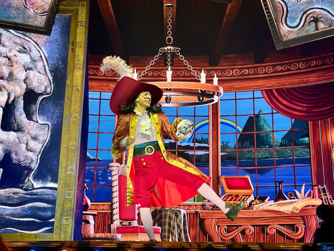 A costumed Captain Hook dances and sings onstage on a pirate ship, with a rainbow and blue sky seen through the windows.