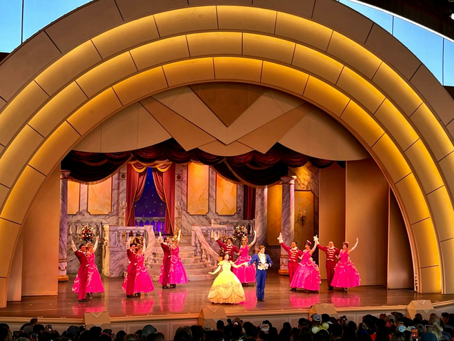 A costumed Belle in a yellow ballgown and the Prince from Disney's 'Beauty and the Beast' dance onstage in a large open-air theater. Behind them are 12 other paired dancers in red ballgowns and suits, dancing.