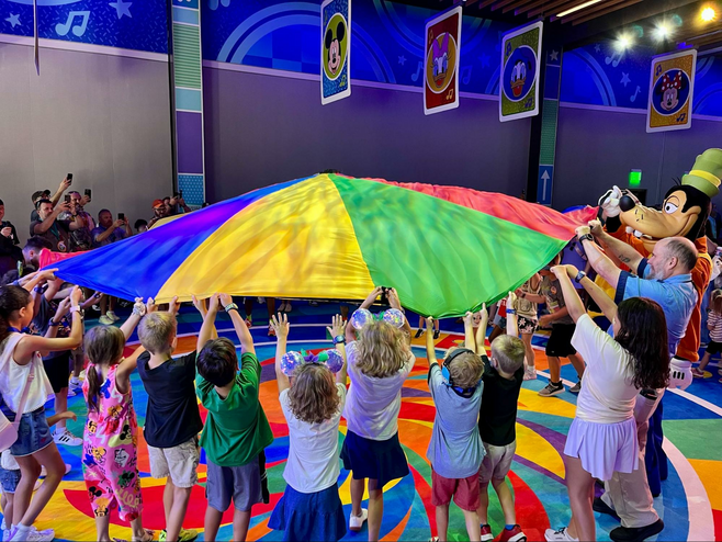 A crowd of young children plays a parachute game on a colorful cushioned dance floor with Goofy.