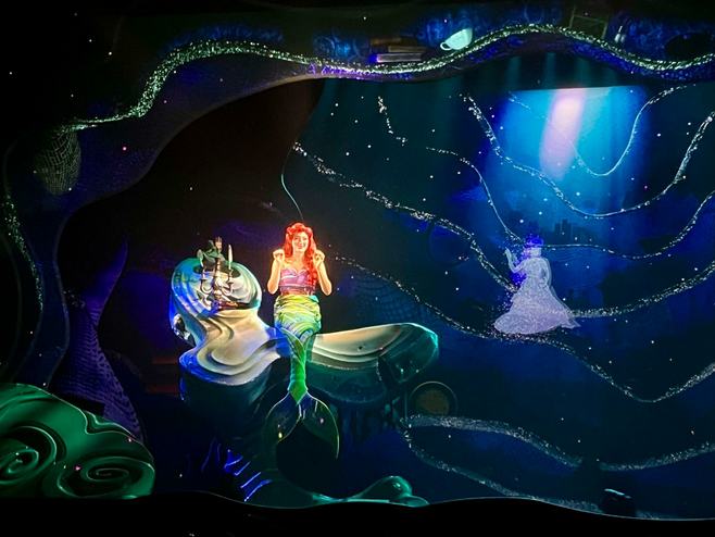Ariel from Disney's The Little Mermaid perches on a rock in her underwater cavern singing to the audience, while a projected image next to her displays a young woman dancing.