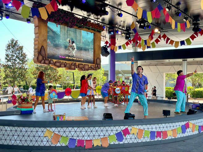 Performers on a small stage sing with audience members to songs from Disney's Encanto as the film plays on a screen behind them.