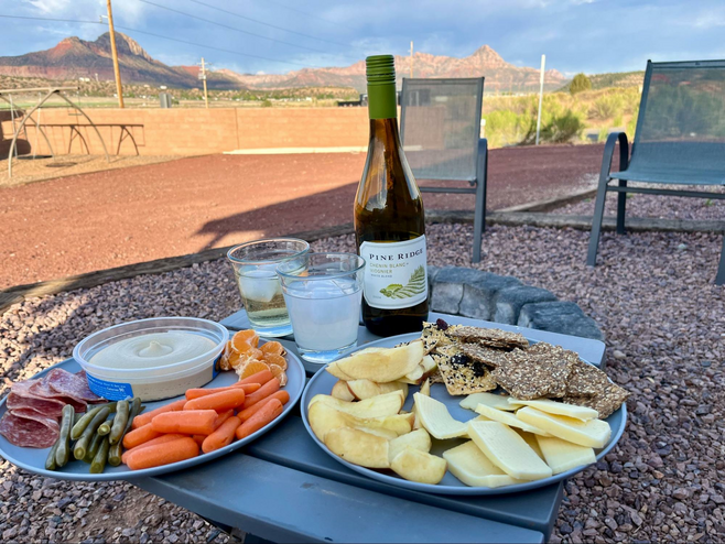 A small table outside holds plates of cheese, salami, fruit, crackers and a wine bottle with two glasses. Mountains in the background.