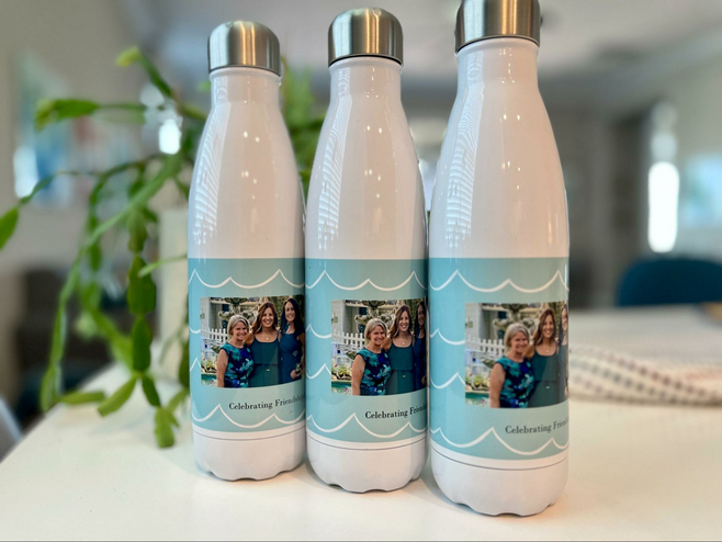 A table holds three white metal water bottles imprinted with photos of three women.