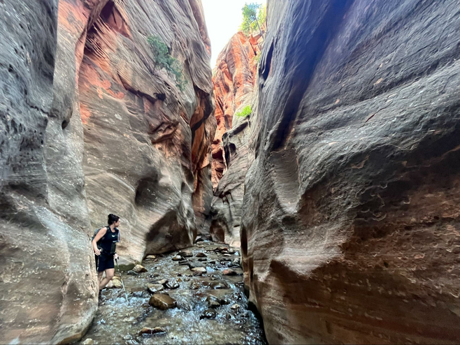 River running through narrow canyon with tall grey and red rock walls surrounding the trail. Hiker walking in the river.