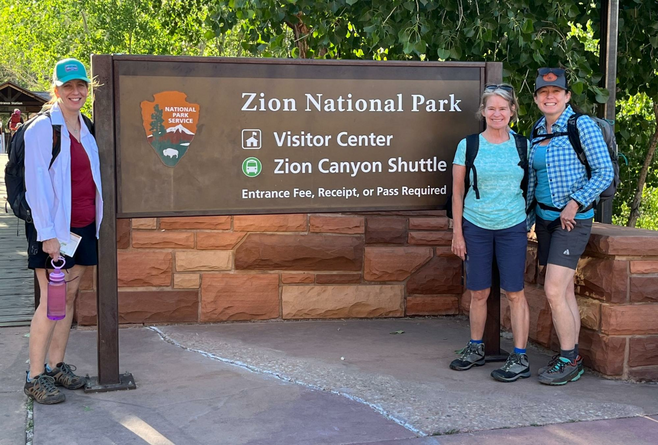 Three female hikers stand next to the Zion National Park entrance sign
