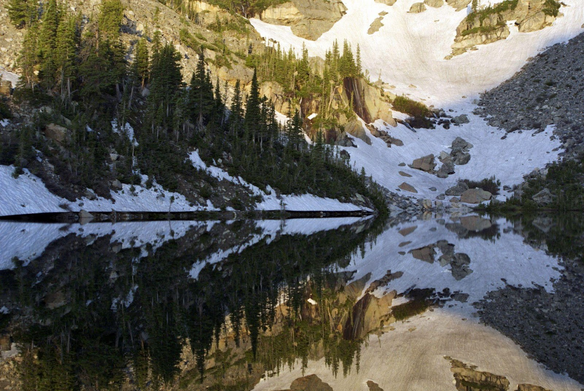 Glass like mountain lake reflecting mountains, forest, and some snow.