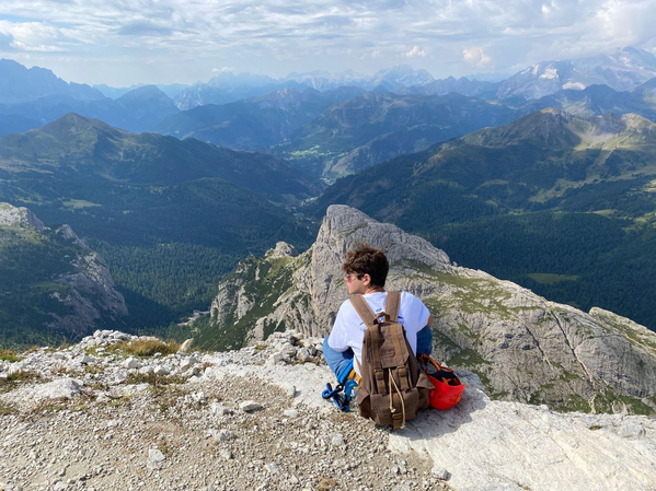 A young man sitting on a ledge overlooking a mountain valley and surrounding peaks.