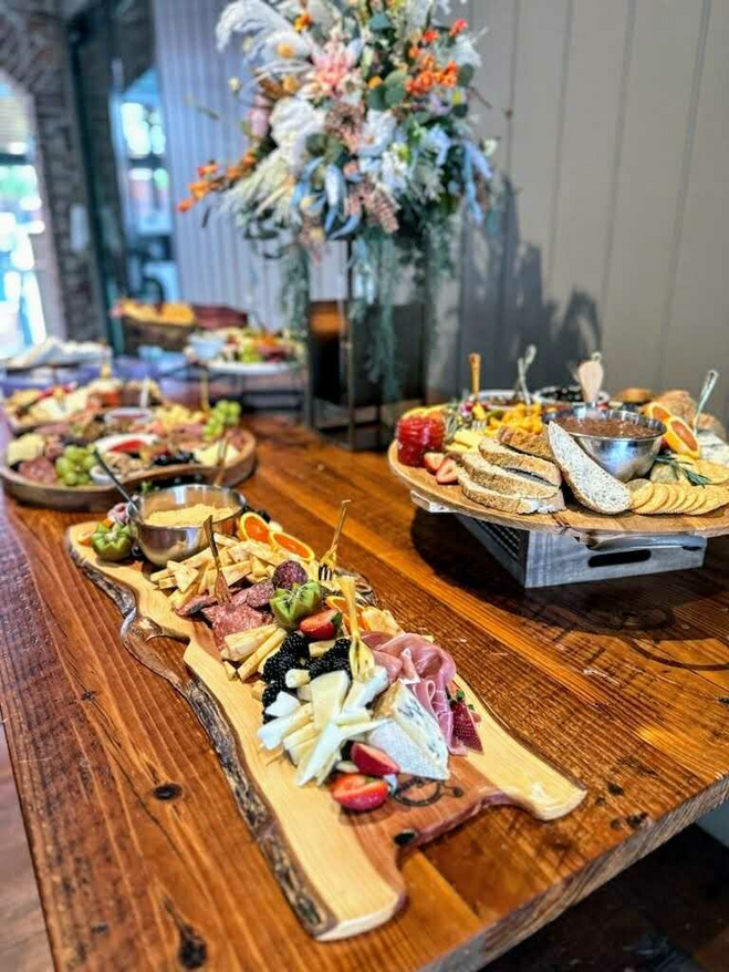 Charcouterie boards with meats, cheeses and fruits on a wooden table with large floral centerpiece.