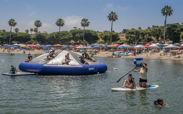 People climbing on the Inflatable water park at Newport Dunes in Newport Beach in front of a crowded beach while swimmers and paddleboarders are nearby.